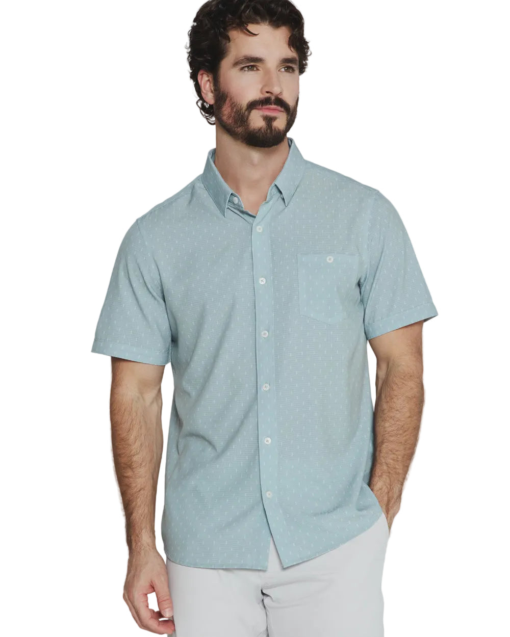A man wearing a light blue short-sleeve button-down shirt with a subtle geometric pattern, paired with white pants, photographed against a light gray studio background.