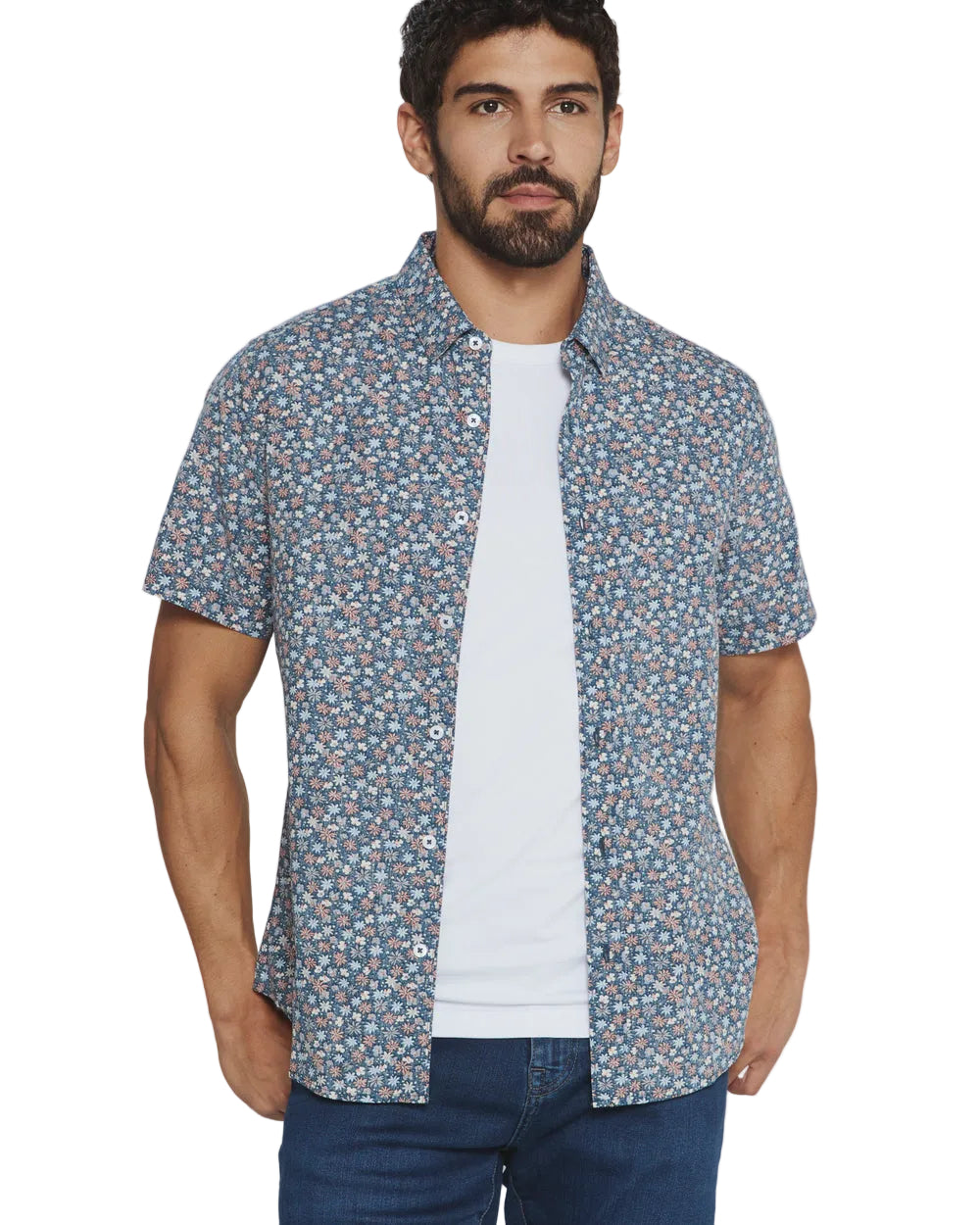 A man wearing a short-sleeve button-up shirt with a dense blue, white, and pink floral pattern, layered over a white t-shirt and blue jeans, against a plain light gray background.